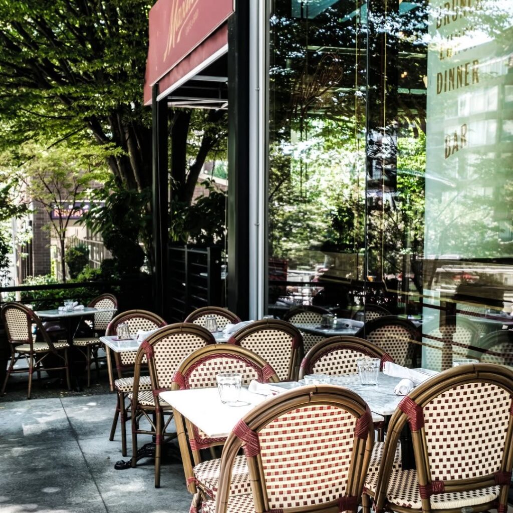 Four-top patio tables by a plate glass window, each surrounded by four wooden chairs with red and white woven seats, under the shade of a large tree